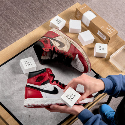 A box of shoe cleaning sponges laid out on a table with 2 hands cleaning a pair of black red and white Nike Air Jordan 1 sneakers. One shoe is dirty and one shoe is clean. Box has text that says "Midsole and Sneaker Cleaner", "Power Pad", "Reshoevn8r".