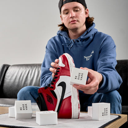 A young male cleaning a black red and white Nike Air Jordan 1 sneaker. Table has numerous shoe cleaning sponges on it. Sponge says "RESHOEVN8R".