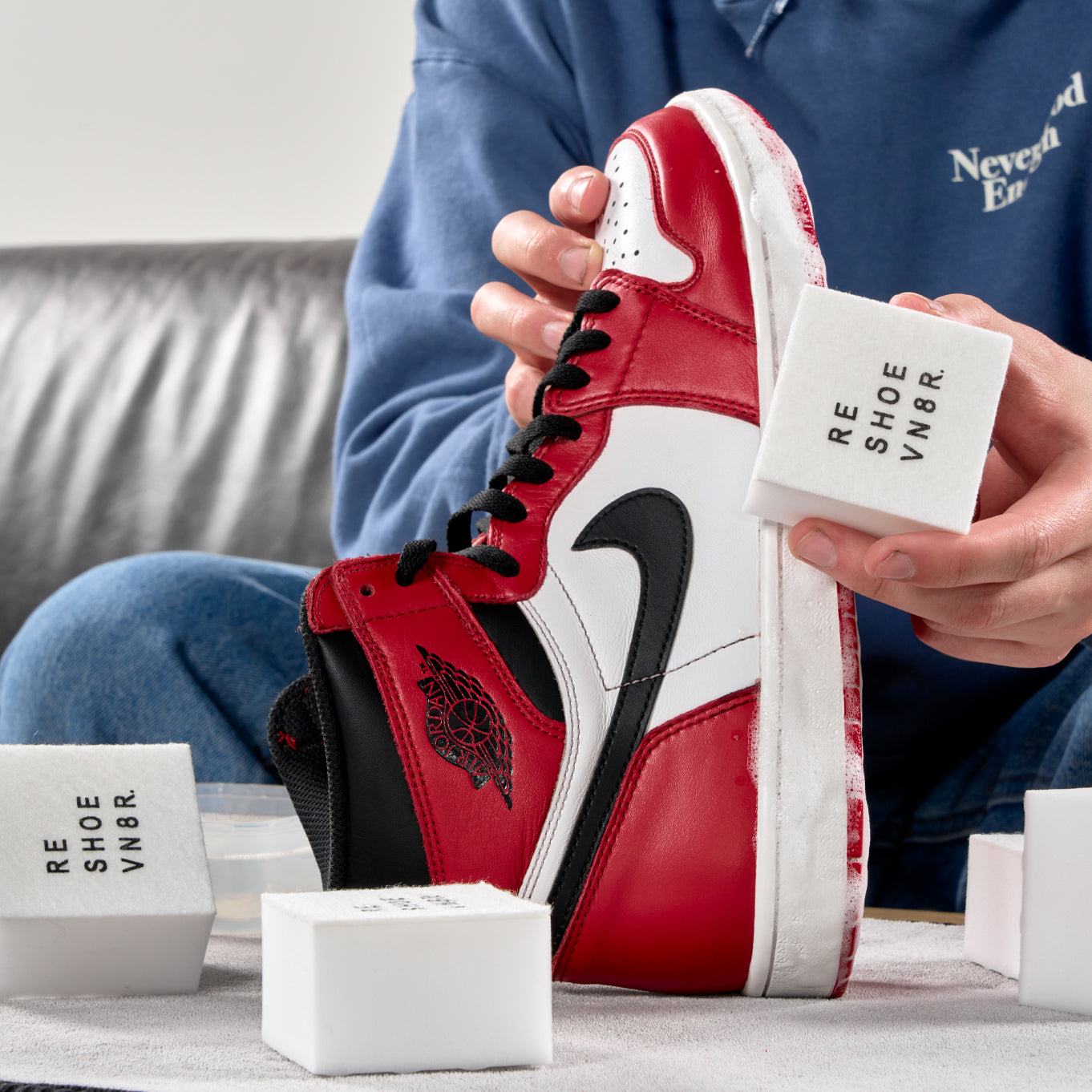 2 hands cleaning a black red and white Nike Air Jordan 1 sneaker. Table has numerous shoe cleaning sponges on it. Sponge says "RESHOEVN8R".