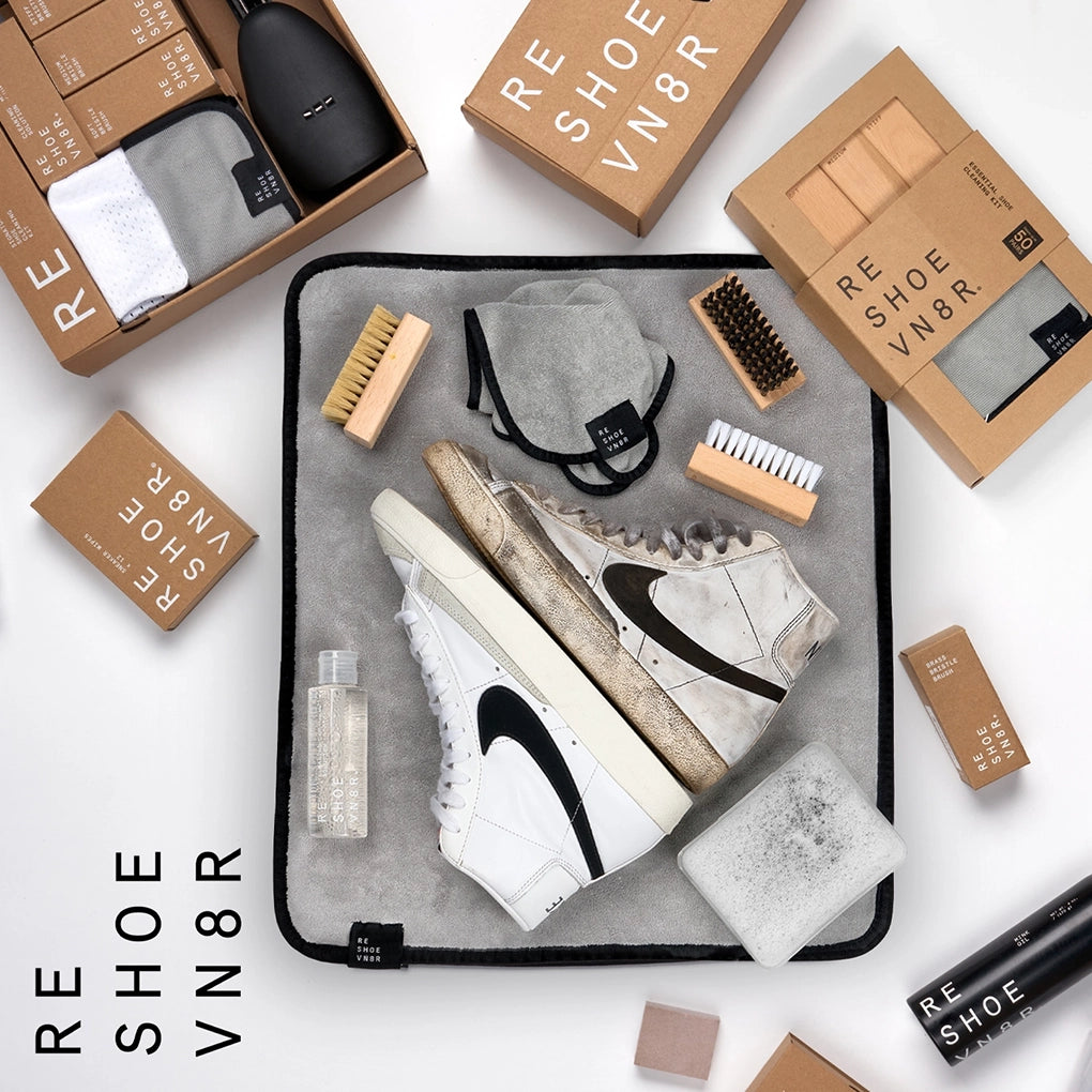 A pair of dirty sneakers, white Nike Blazers, sit kicked back next to a box of Reshoevn8r shoe cleaning supplies on a light colored floor. The cleaning supplies include a bottle of cleaning solution, three different bristle brushes, and a microfiber towel. Text on the box indicates that the kit is suitable for suede, leather, mesh, canvas, nubuck, plastic and rubber shoes.